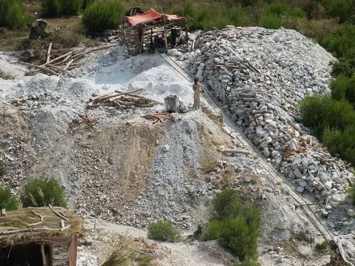 Hillside mine site with waste dumps, a chute and a small shed among green vegetation.