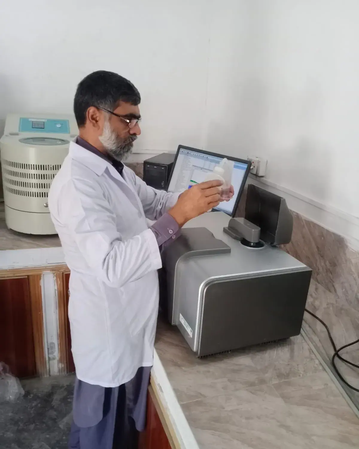 Laboratory technician in a white coat operating an analytical instrument connected to a computer.